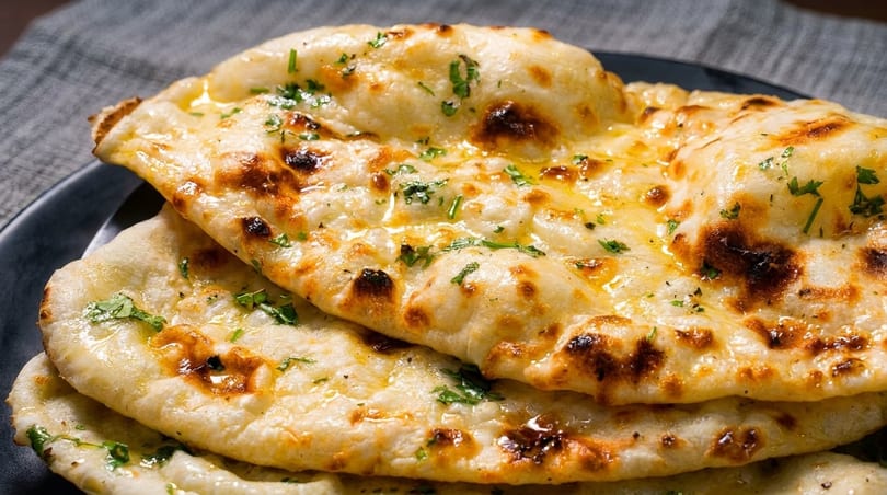 Golden-brown naan bread, brushed with butter and sprinkled with fresh herbs, stacked on a dark plate.