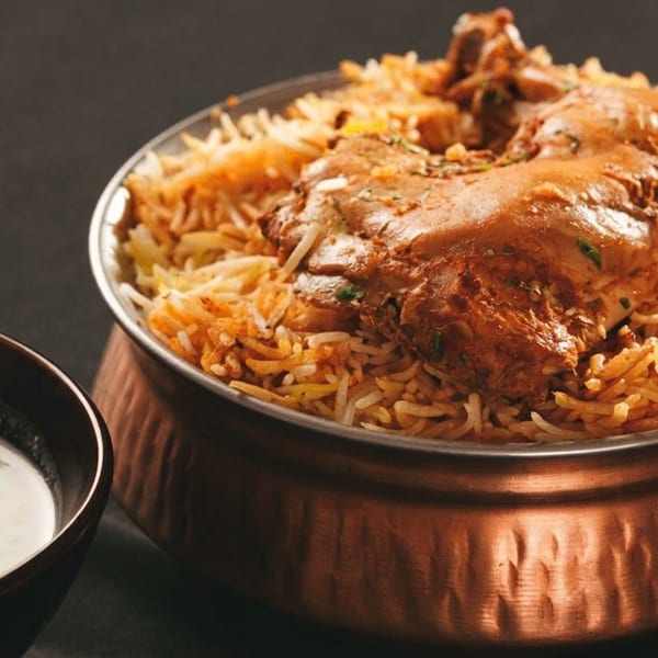 Chicken biryani in a copper pot with a side of raita, on a dark surface.