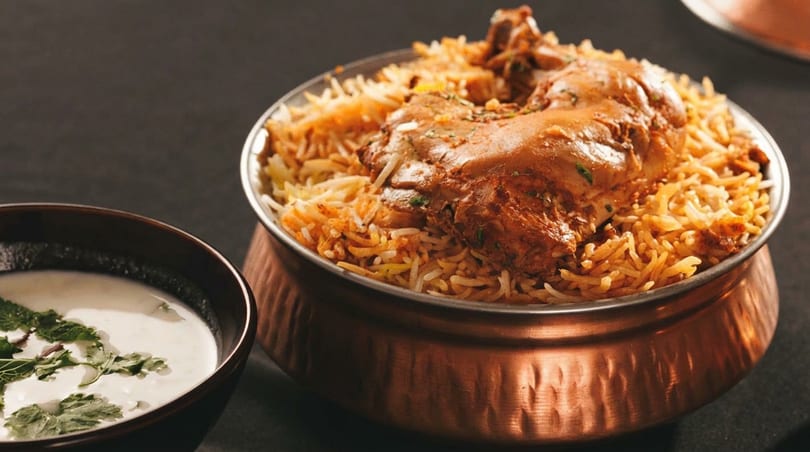 Chicken biryani in a copper pot with a side of raita, on a dark surface.