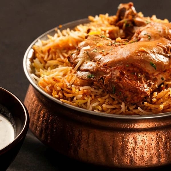 Chicken biryani in a copper pot with a side of raita, on a dark background.