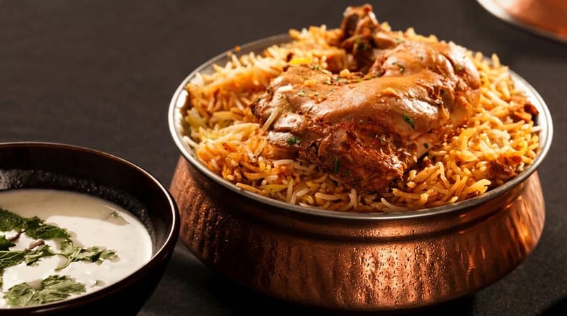 Chicken biryani in a copper pot with a side of raita, on a dark background.