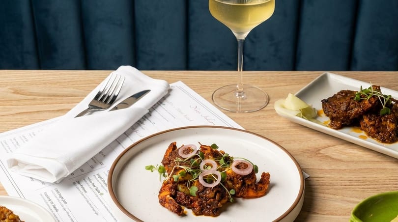 Indian dishes of shrimp and meat, a glass of white wine, and a menu on a wooden table.