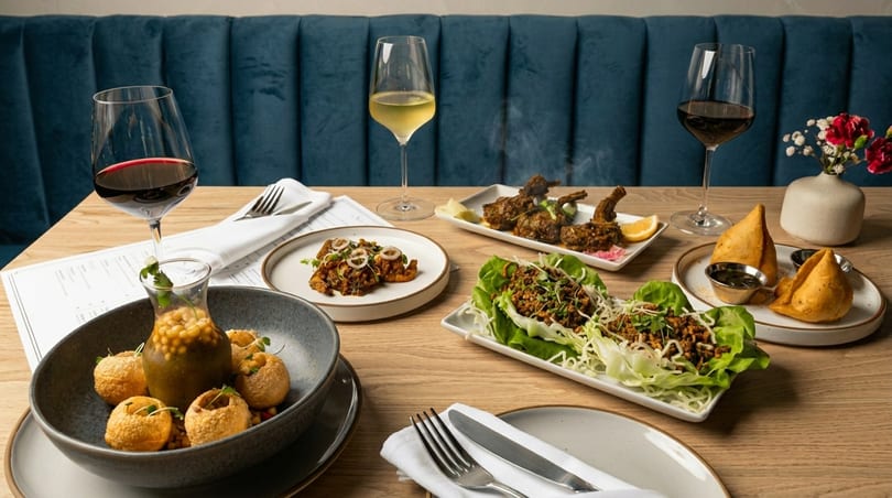 A table laden with various Indian dishes, including samosas, pani puri, and lamb chops, with wine glasses.