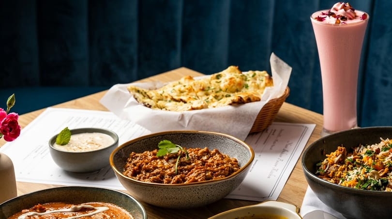 Indian dishes, including naan, curries, rice, and a pink lassi, on a wooden table.