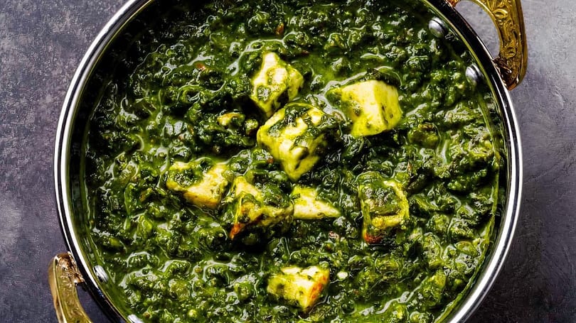 Close-up of Palak Paneer, a vibrant green spinach curry with cubes of paneer cheese in a metal bowl.