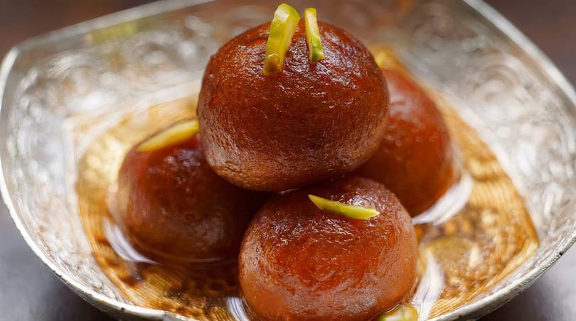 Four glistening gulab jamun, garnished with slivered pistachios, in a decorative silver bowl.
