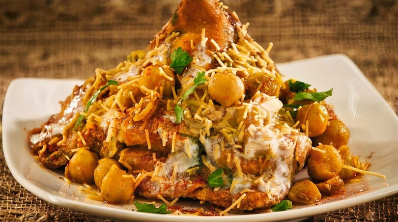 A close-up of a samosa chaat, topped with chickpeas, yogurt, chutneys, and crispy noodles.