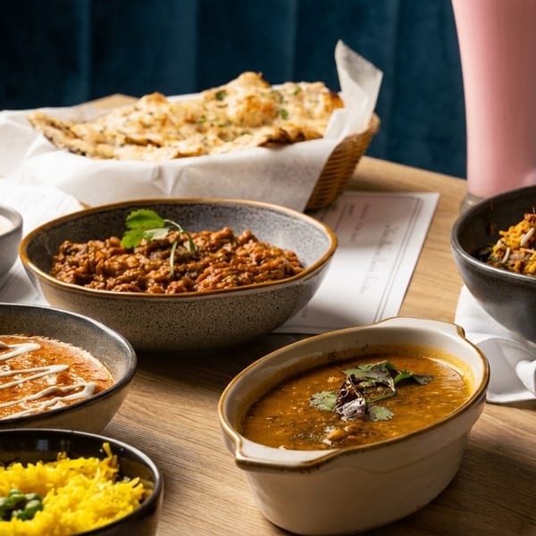 A table laden with various Indian dishes including curries, rice, naan bread, and a pink drink.