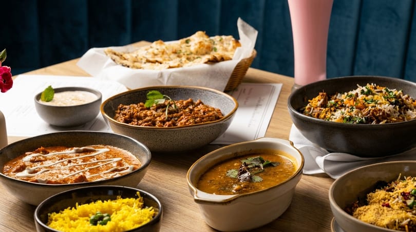 A table laden with various Indian dishes including curries, rice, naan bread, and a pink drink.