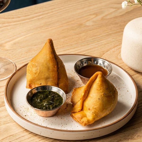 Two golden-brown samosas with green and brown dipping sauces, a glass of red wine, and a small vase of flowers on a wooden table.