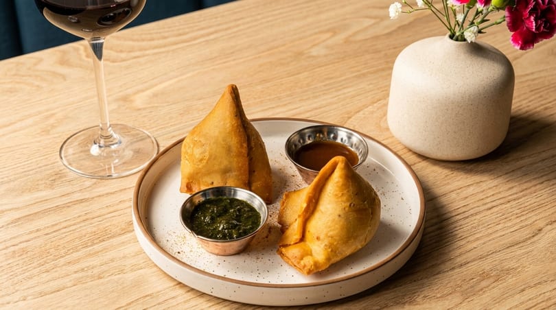 Two golden-brown samosas with green and brown dipping sauces, a glass of red wine, and a vase of flowers.
