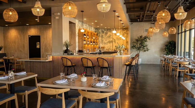 A modern restaurant interior with a bar, dining tables, woven light fixtures, and botanical wallpaper.